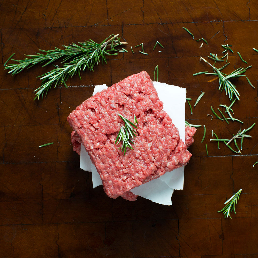 Stack of ground veal meat on a paper, garnished with a sprig of rosemary, on a wooden surface with scattered rosemary leaves.