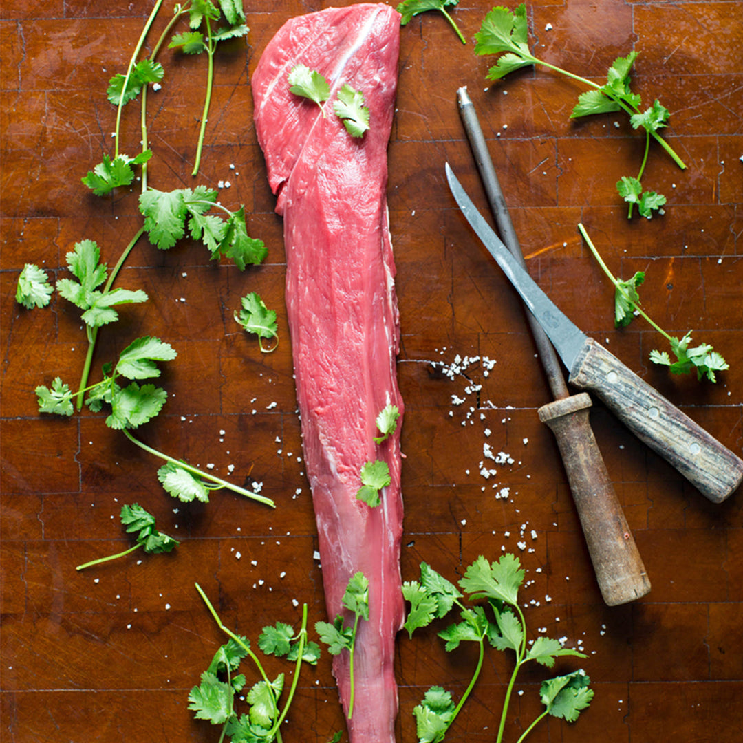 A whole Kentucky Grass-Fed Rose Veal Tenderloin on a wooden surface with herbs and spices scattered around, along with a knife and fork.